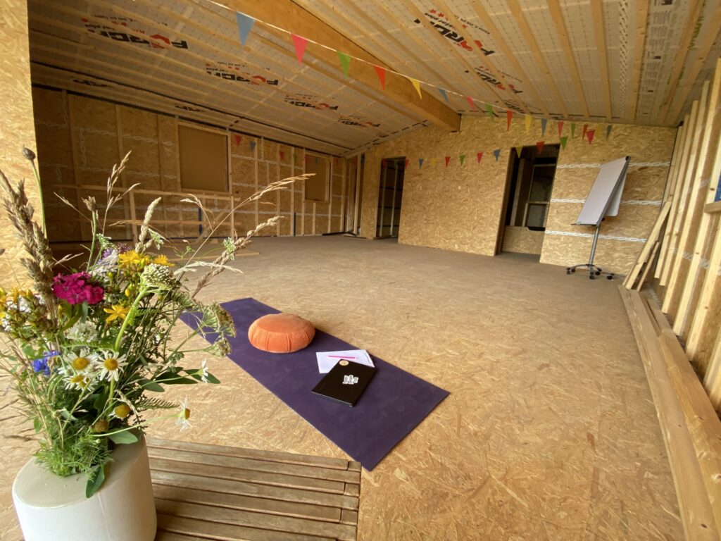 Eine Yogamatte mit Meditationskissen wurde im Rohbau eines Workshopraumes ausgerollt. Daneben steht ein Blumenstrauß.